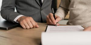 Dwóch mężczyzn przy biurku podczas podpisywania dokumentów, jeden trzyma długopis, drugi nadzoruje przebieg formalności