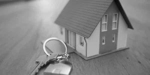 Model małego domu stojący na drewnianym blacie obok pęku kluczy z brelokiem w kształcie domu.