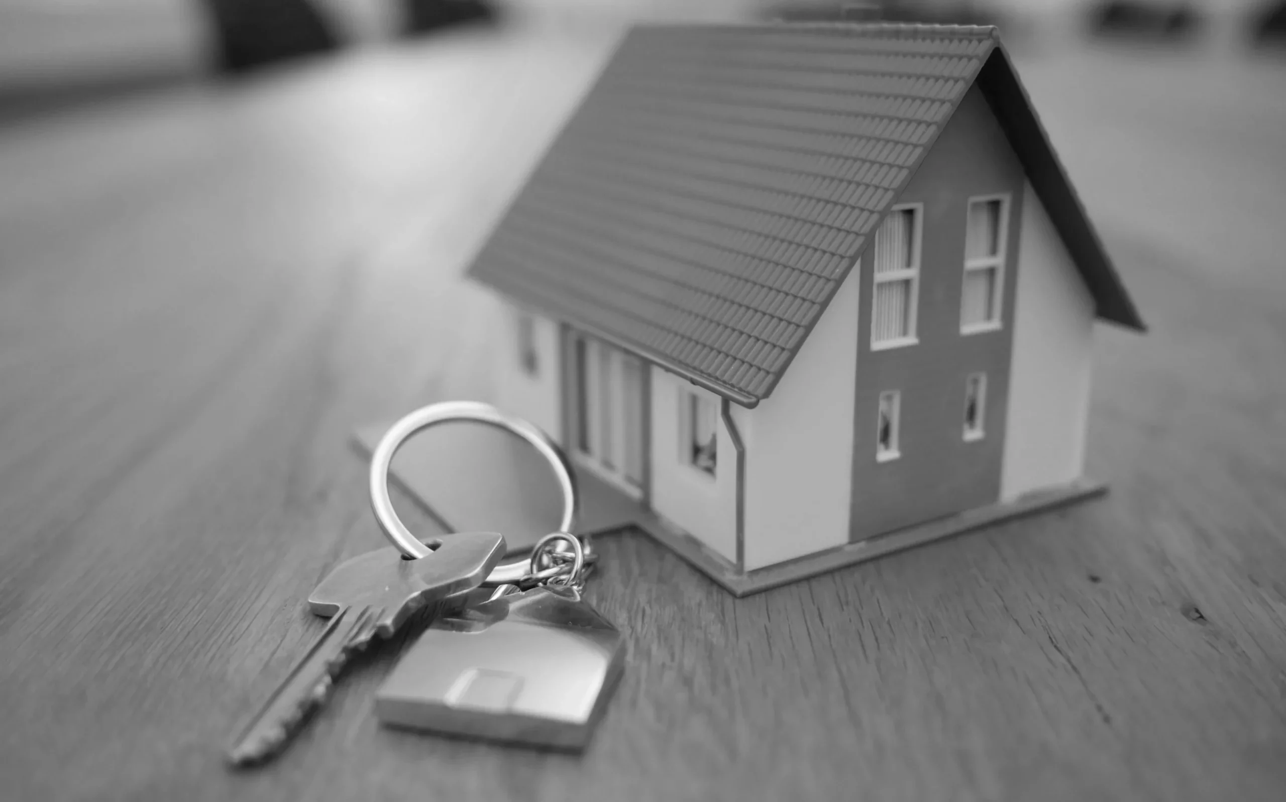 Model małego domu stojący na drewnianym blacie obok pęku kluczy z brelokiem w kształcie domu.